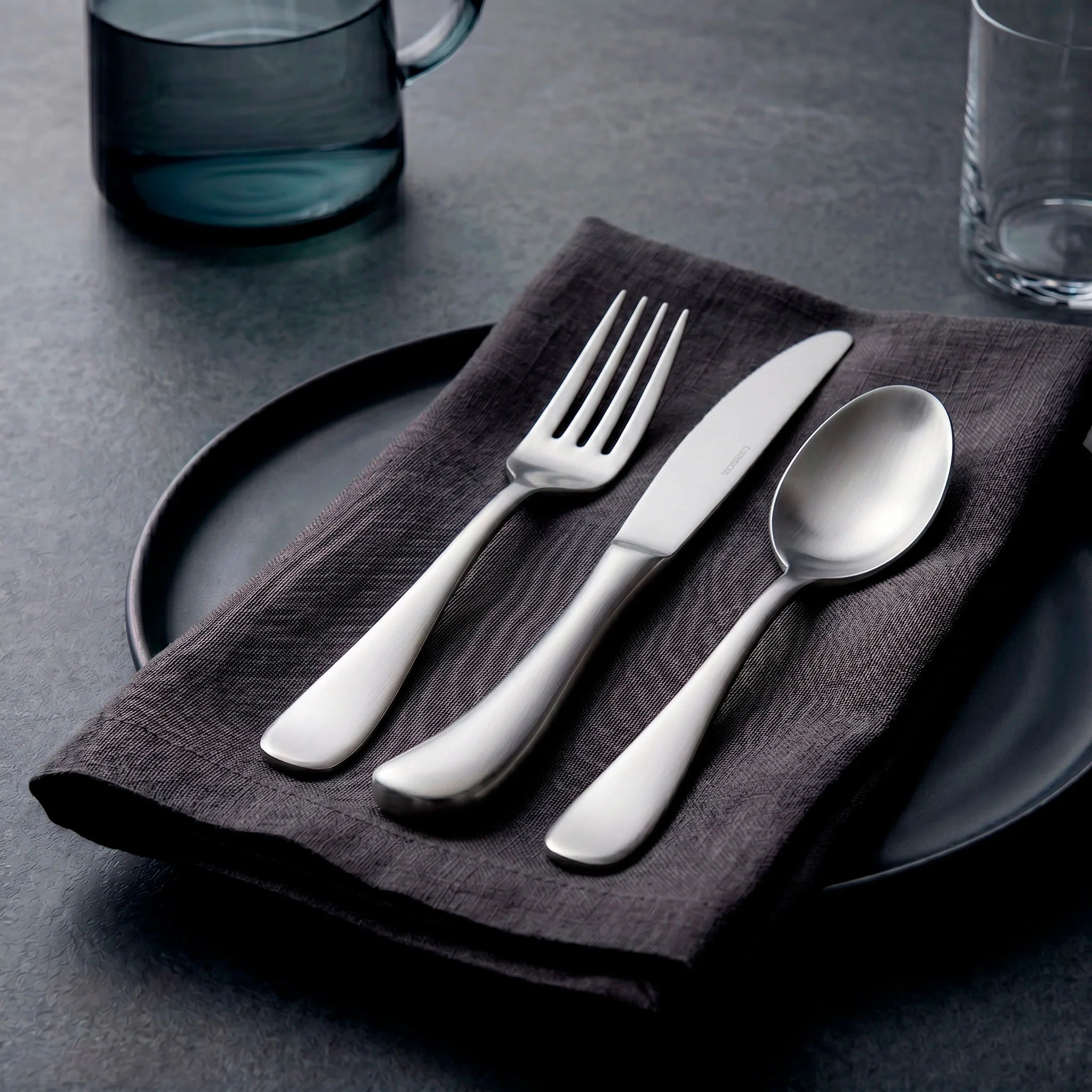 Set de cubiertos premium de la colección Mariposa Brushed de Fortessa con acabado mate cepillado sobre una servilleta gris de lino